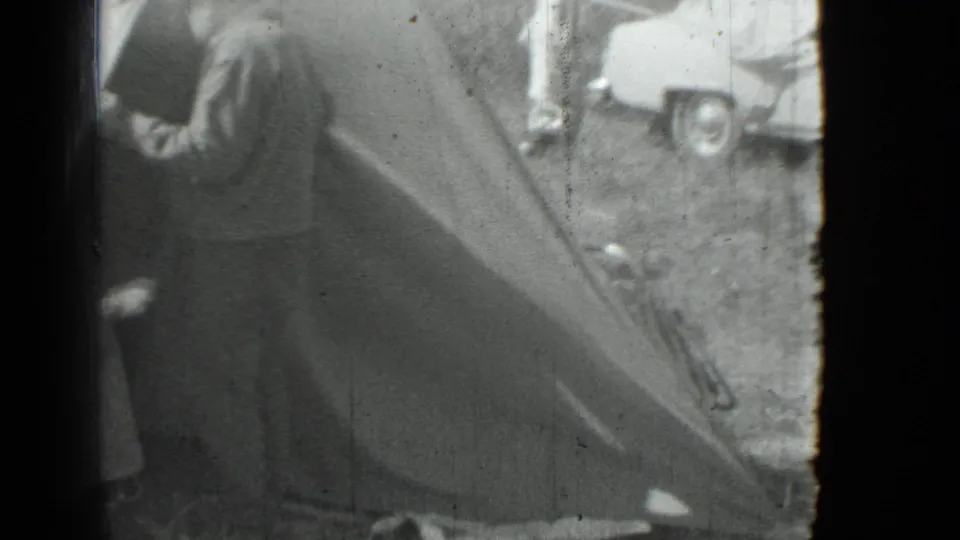 Black-and-white 1967 home-movie frame showing a man working near a large tent, with part of a car visible behind him.