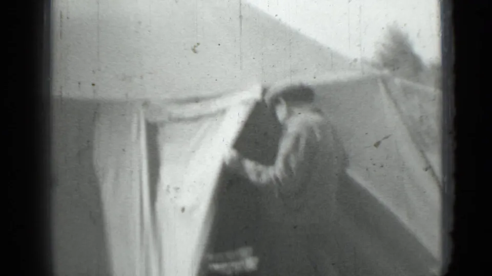 Black-and-white home-movie frame from the supplied 1967 clip showing a woman standing or walking beside a large tent in a field.