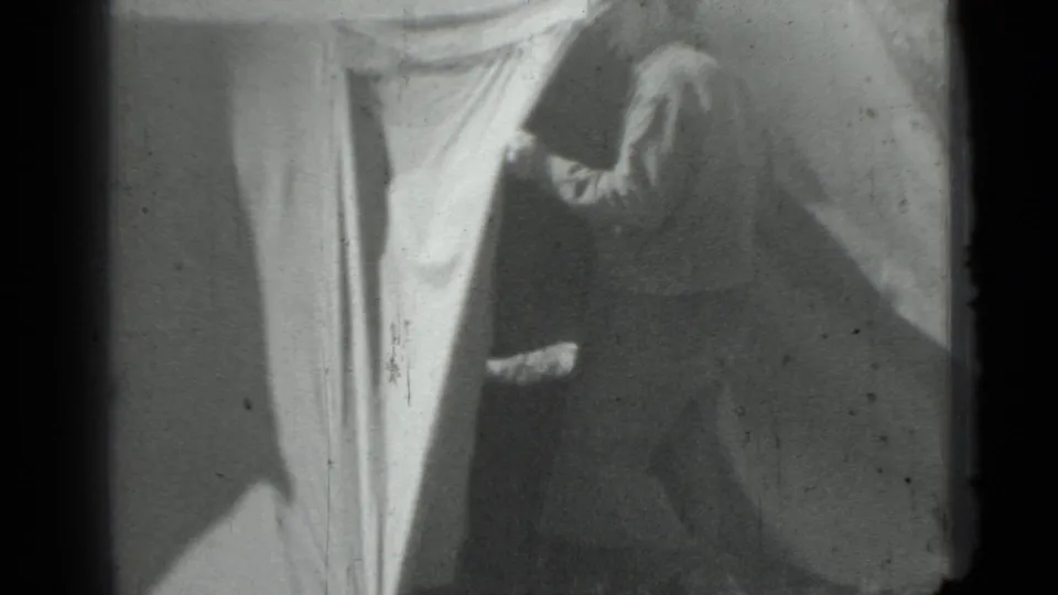 Black-and-white 8mm frame showing a woman near the entrance or side of a large tent, with canvas fabric dominating the view.