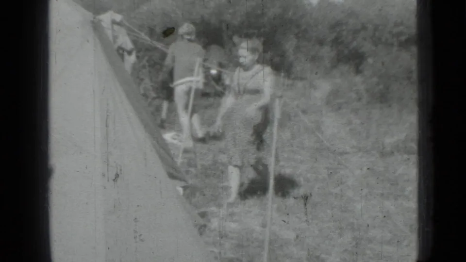 Black-and-white 1967 home-movie frame showing two adults near a tent in an open grassy area, with background objects partly visible.