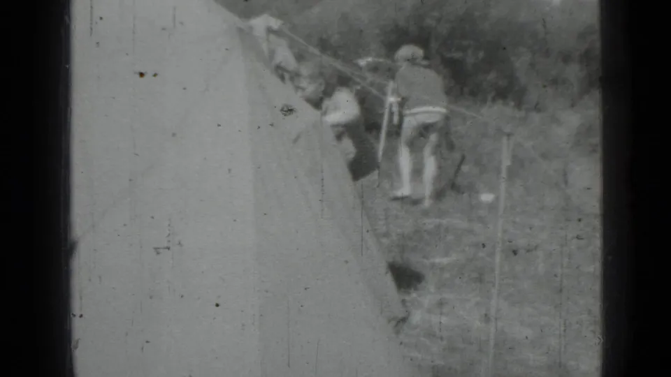 A black-and-white 8mm frame shows two women working around guy lines near a pitched canvas tent at the campsite.
