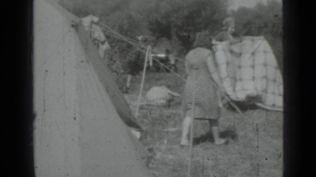 Two women stand by hanging laundry near a tent and clothesline in a grassy outdoor area.