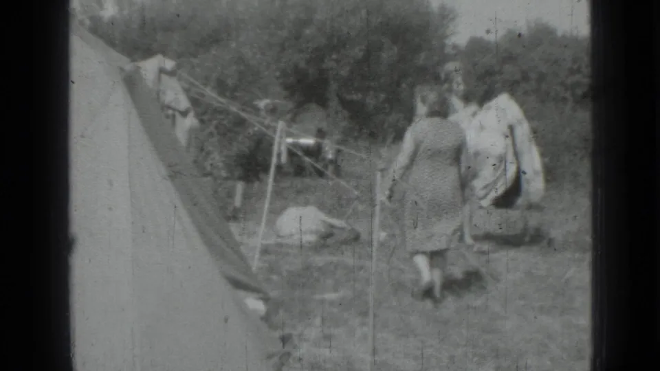 Two adult women walk through a grassy campsite beside a canvas tent, with clotheslines and trees visible in the 1967 home-movie frame.