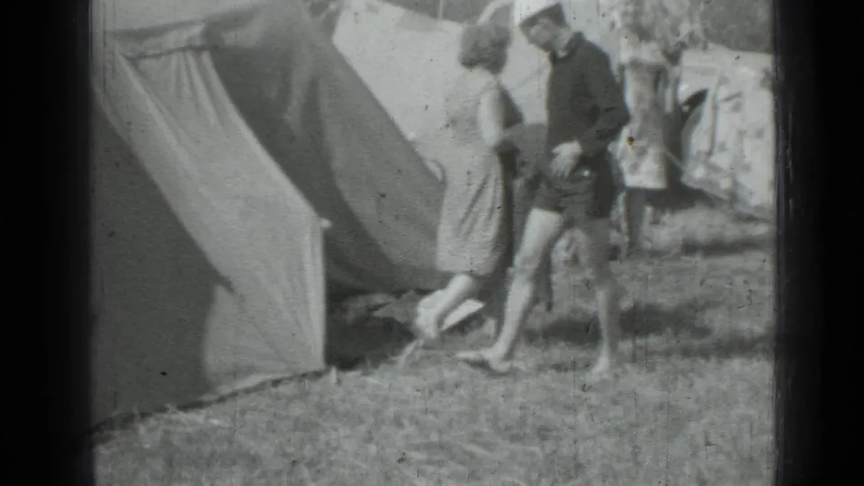1967 home-movie frame from Saint Petersburg, Russia, showing adults moving beside a canvas tent at a grassy campsite.
