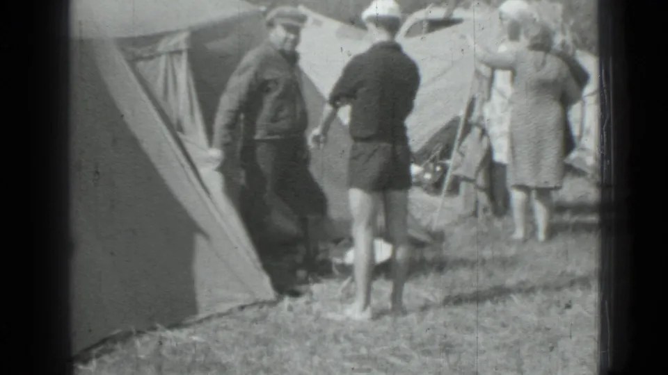 At the campsite, a man in shorts stands near another adult by the tent while a woman in a dress faces away.