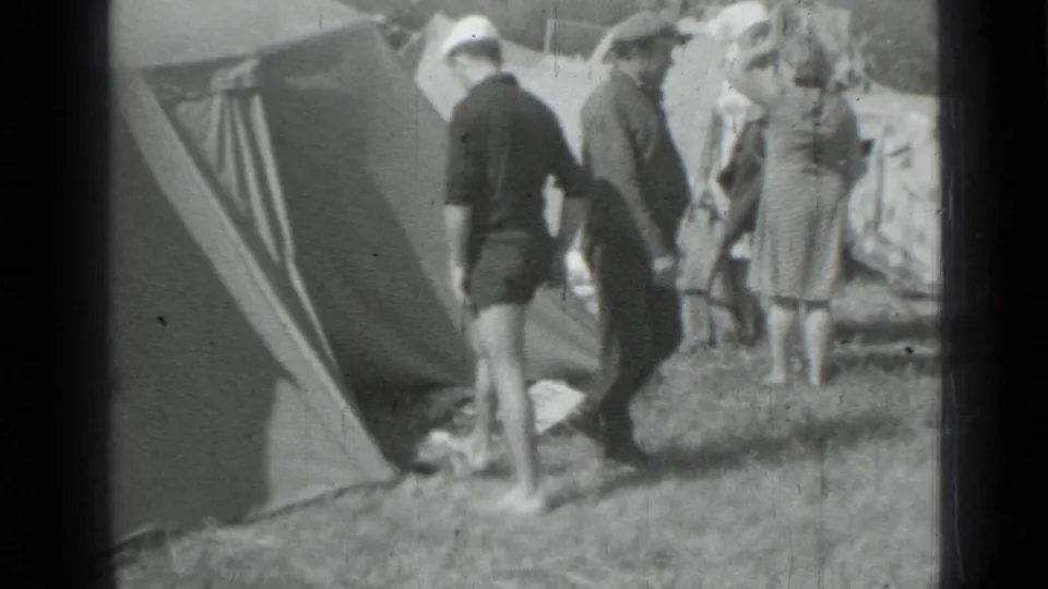Adults stand and converse beside a canvas tent at a grassy 1967 Saint Petersburg campsite, with camp items at their feet.