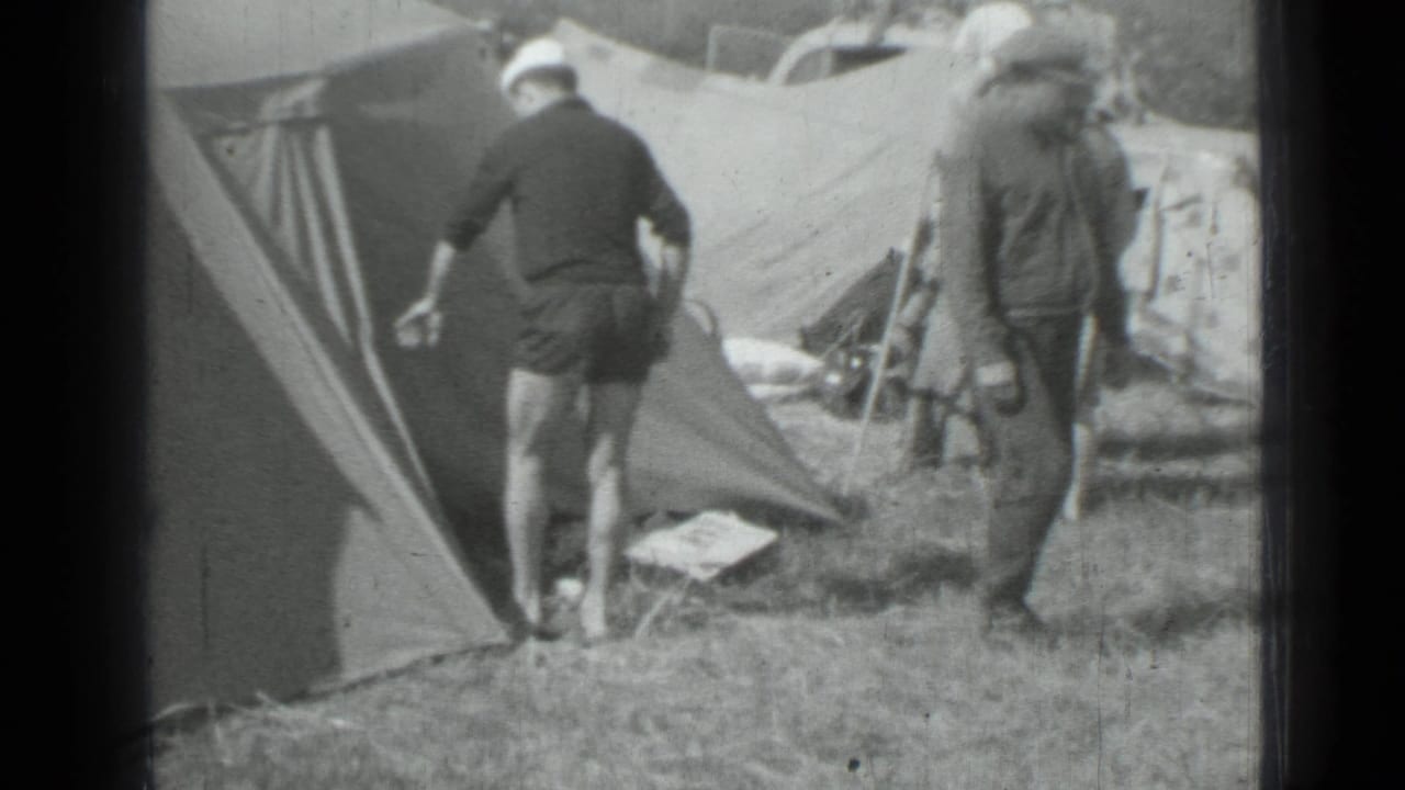 Adults stand beside canvas tents on a grassy campsite.