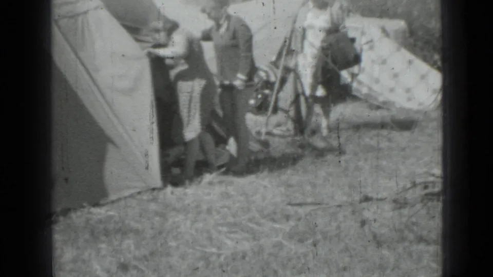 1967 home-movie frame showing adults gathered beside canvas tents and gear at a grassy campsite.