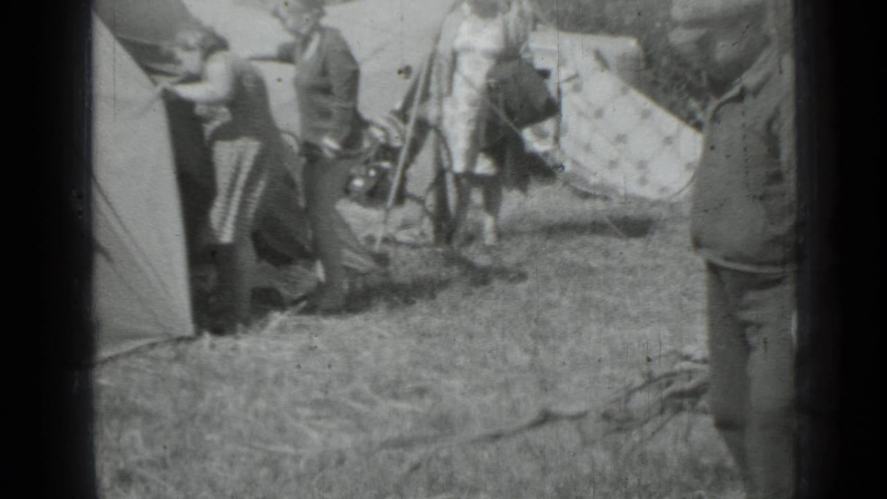 Adults stand beside canvas tents in a grassy campsite with camping gear nearby.