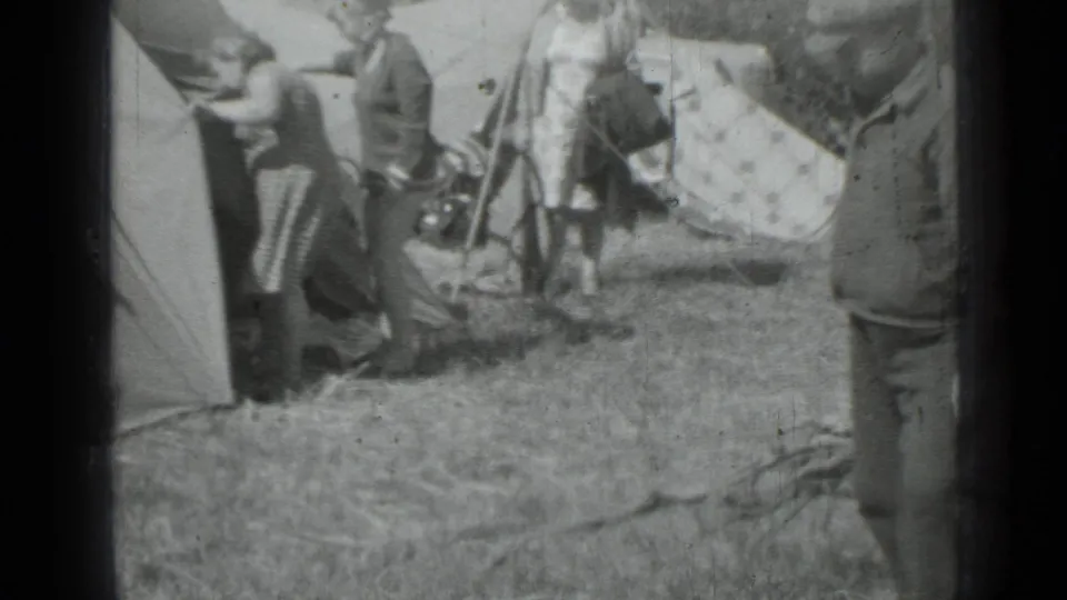 1967 8mm home-movie frame of adults around canvas tents in a grassy campsite, with a bicycle and gear partly visible.