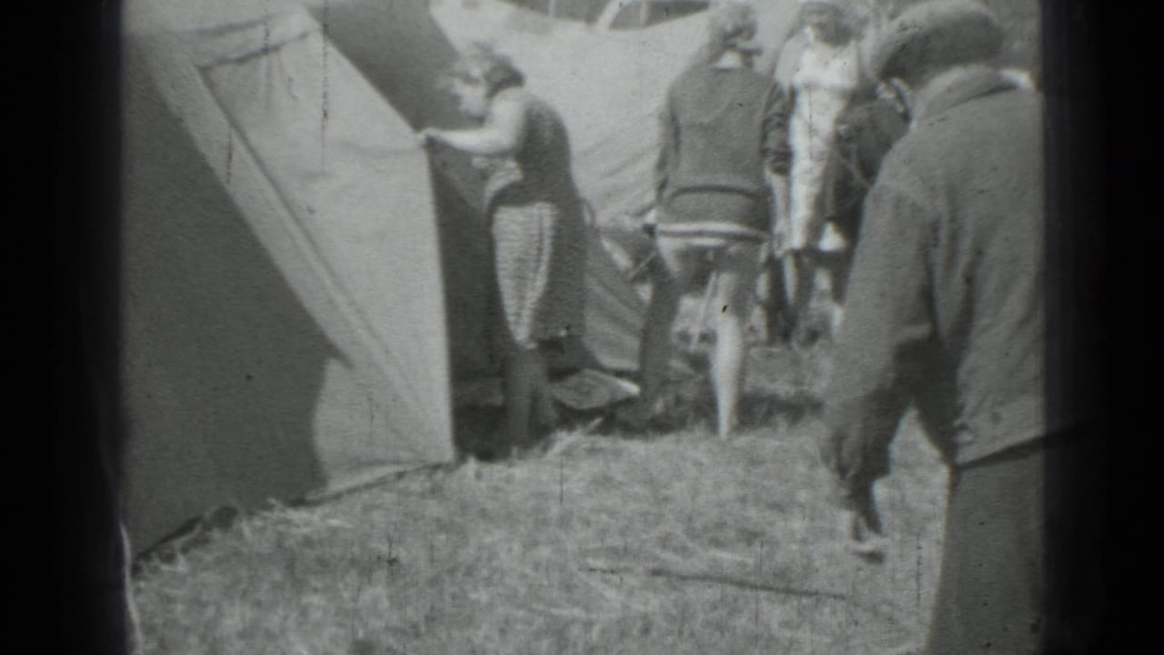 Adults stand near a tent while a dog moves at the edge of the campsite.