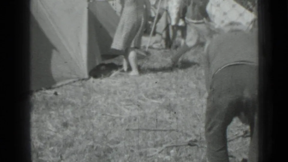 Black-and-white 8mm frame from a 1967 camping scene, with adults near tent fabric and a person bending in the foreground.