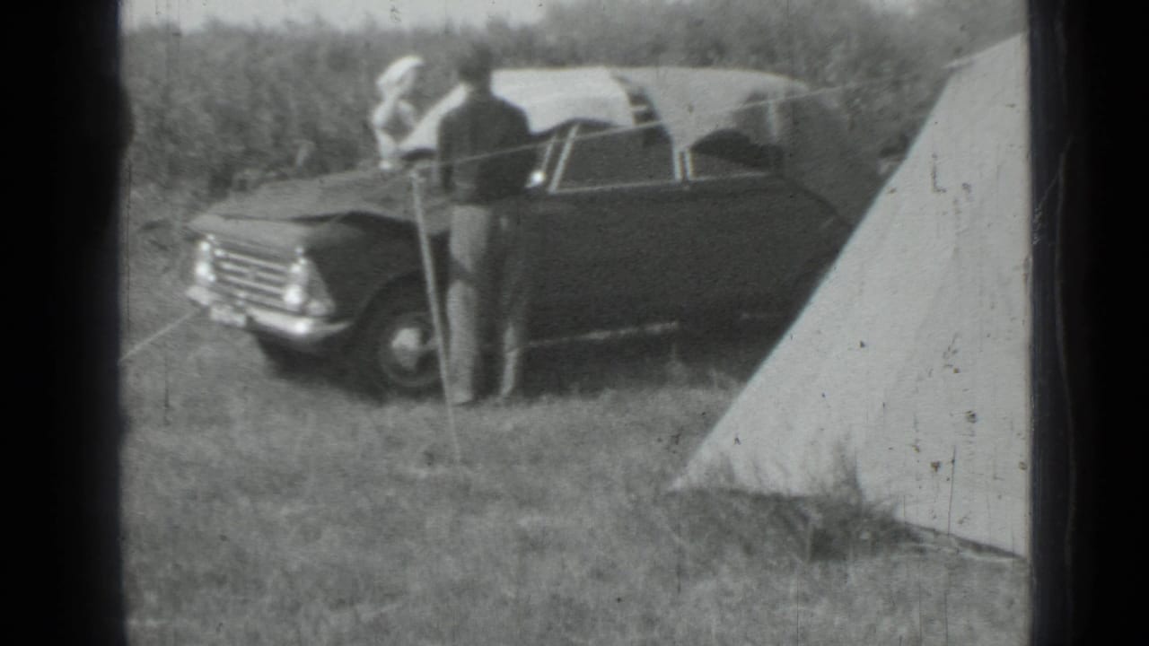Dark car parked beside canvas tents in a grassy camp area with an adult standing nearby.