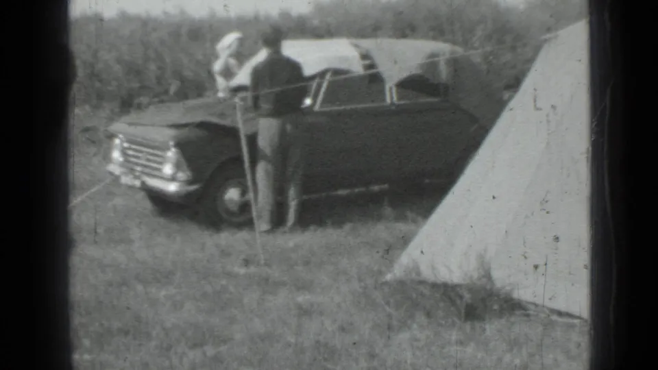 1967 Saint Petersburg home-movie frame showing a dark period car, canvas tents, and an adult standing at a grassy campsite.