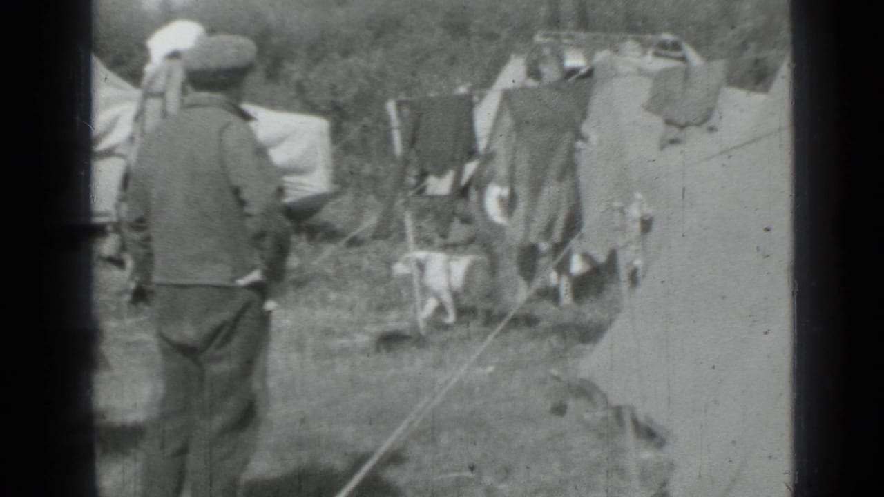 Adults stand near canvas tents and a clothesline at a grassy campsite.