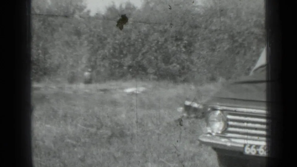 1967 Saint Petersburg home-movie frame showing a sedan front corner near a grassy field and tree line.