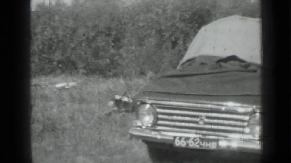 1967 Saint Petersburg frame showing a tarp-covered sedan parked in a grassy field with trees behind it.