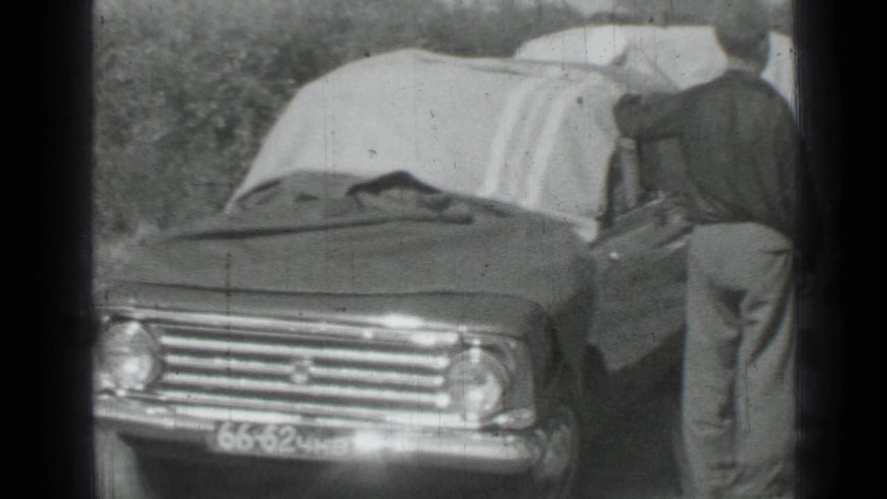 1967 Saint Petersburg home-movie frame showing a man beside a tarp-covered sedan in a grassy field.