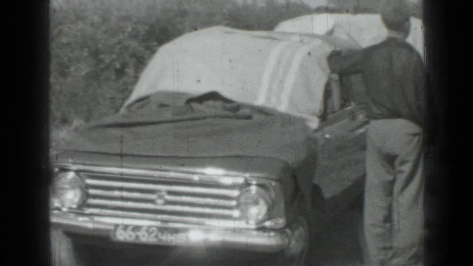 1967 Saint Petersburg home-movie frame showing a man beside a tarp-covered sedan in a grassy field.