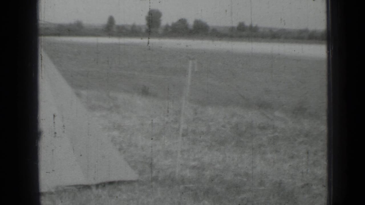 Monochrome 8mm frame showing a rural field, pale tarp edge, posts, and a distant tree line in the reported Saint Petersburg area.