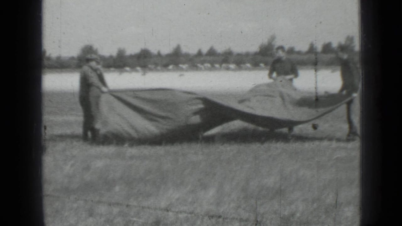 1967 black-and-white home-movie frame showing figures in a field spreading or holding a large tarp near open water or a pale road.