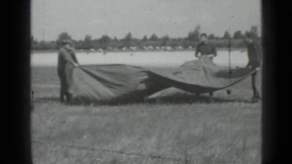 1967 black-and-white home-movie frame showing figures in a field spreading or holding a large tarp near open water or a pale road.