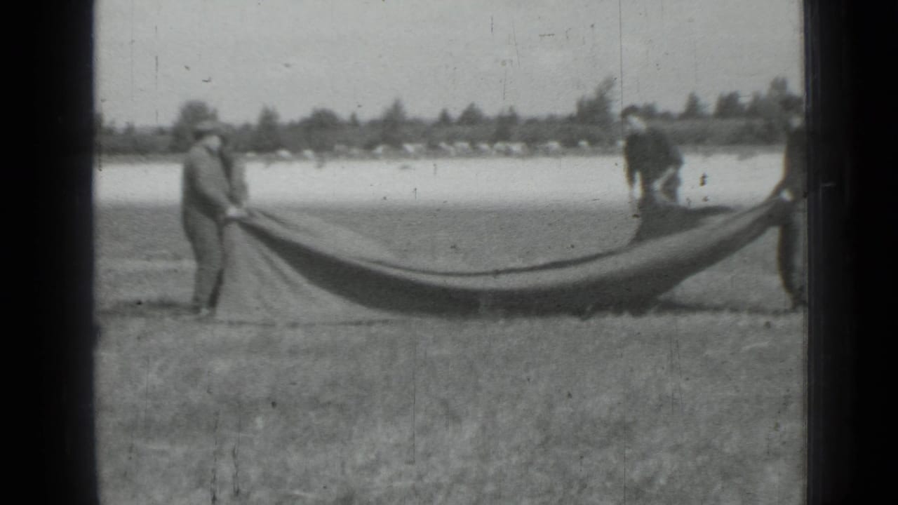 In supplied 1967 Saint Petersburg-area 8mm footage, men hold a large dark fabric across a grassy field with trees in the distance.
