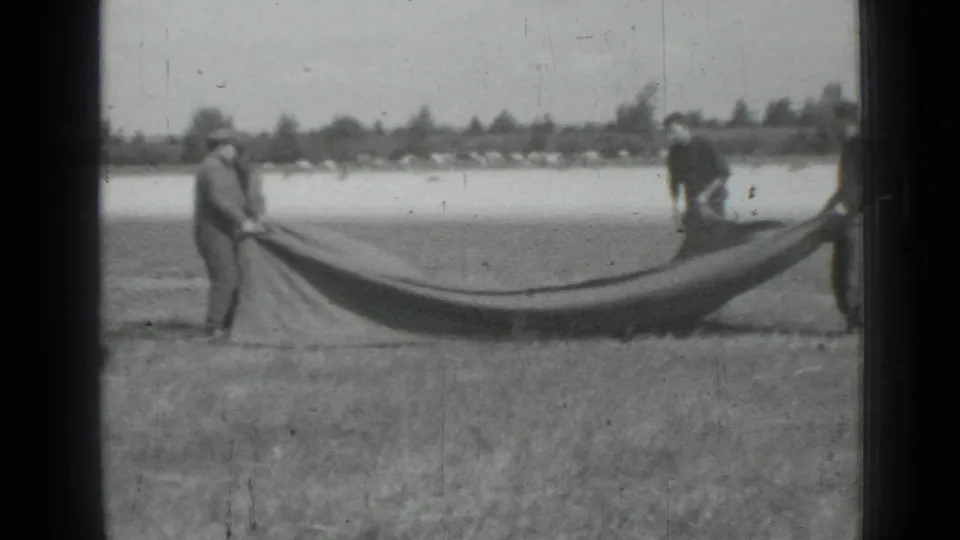 In supplied 1967 Saint Petersburg-area 8mm footage, men hold a large dark fabric across a grassy field with trees in the distance.