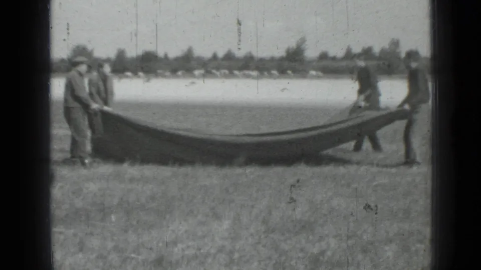 Several men in vintage clothing hold a sagging blanket or tarp in an open field, with a pale waterline or background beyond.