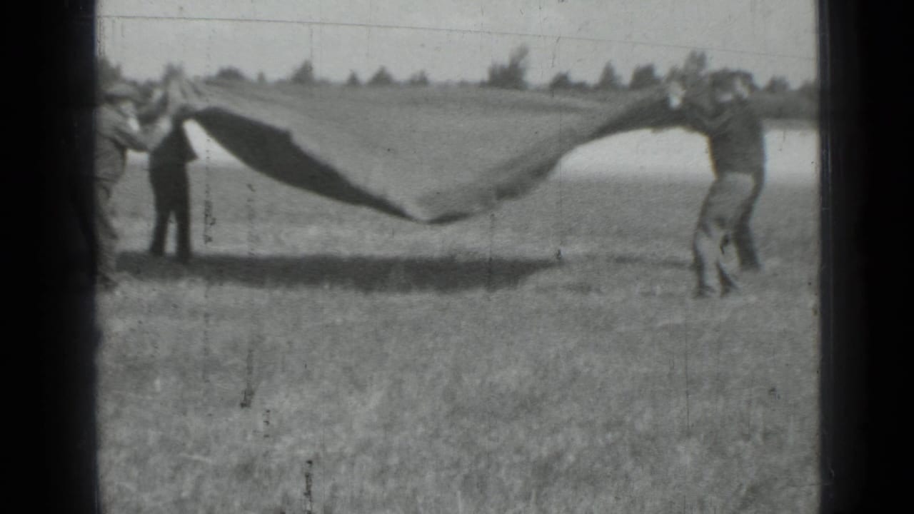 The men lift or shake the large dark fabric, which rises and billows above the grassy ground in the home-movie frame.