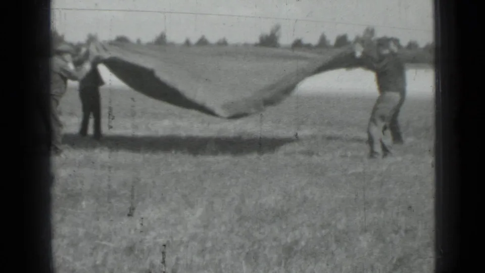 The men lift or shake the large dark fabric, which rises and billows above the grassy ground in the home-movie frame.
