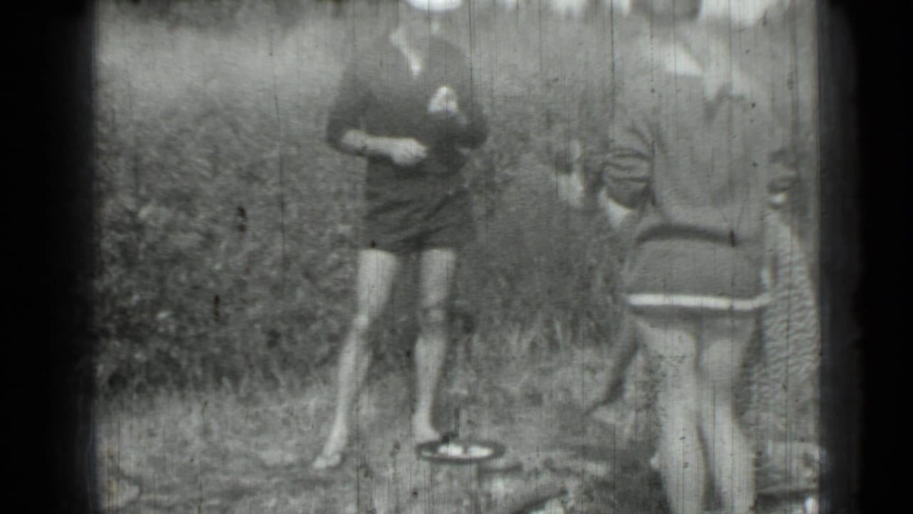 Grainy 1967 8mm frame showing a woman in a dark dress beside a pan while another woman stands nearby.