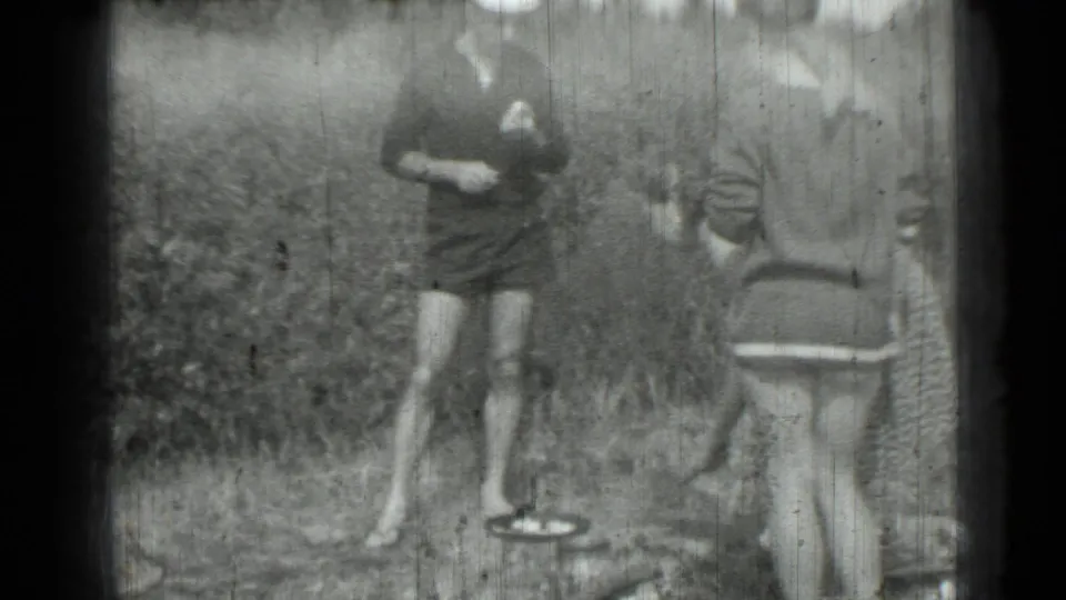 Grainy 1967 8mm frame showing a woman in a dark dress beside a pan while another woman stands nearby.