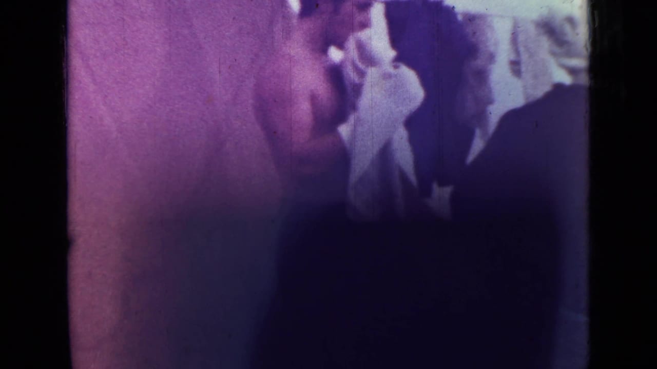 Close, soft 8mm view of a shirtless man and white cloth beside another figure, with purple color shift and visible film scratches.