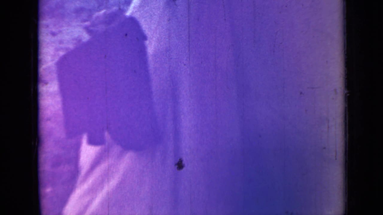 Blurred 1967 8mm frame shows a figure near bright tent-like fabric, with strong purple fading and heavy grain.