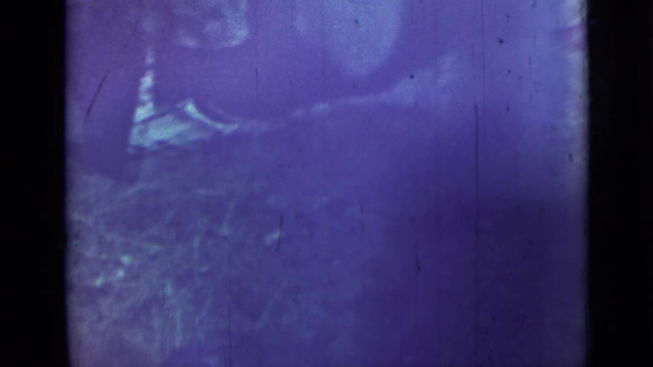 Purple-blue frame with a person bending near a low outdoor area, partly obscured by film fading.