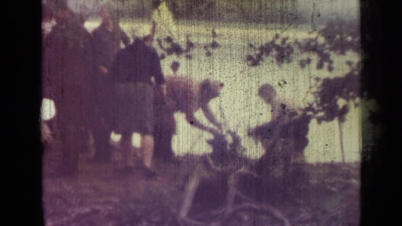 1967 8mm home-movie frame near Saint Petersburg showing adults beside a lake, with a tent, shoreline, and overhanging branches visible.