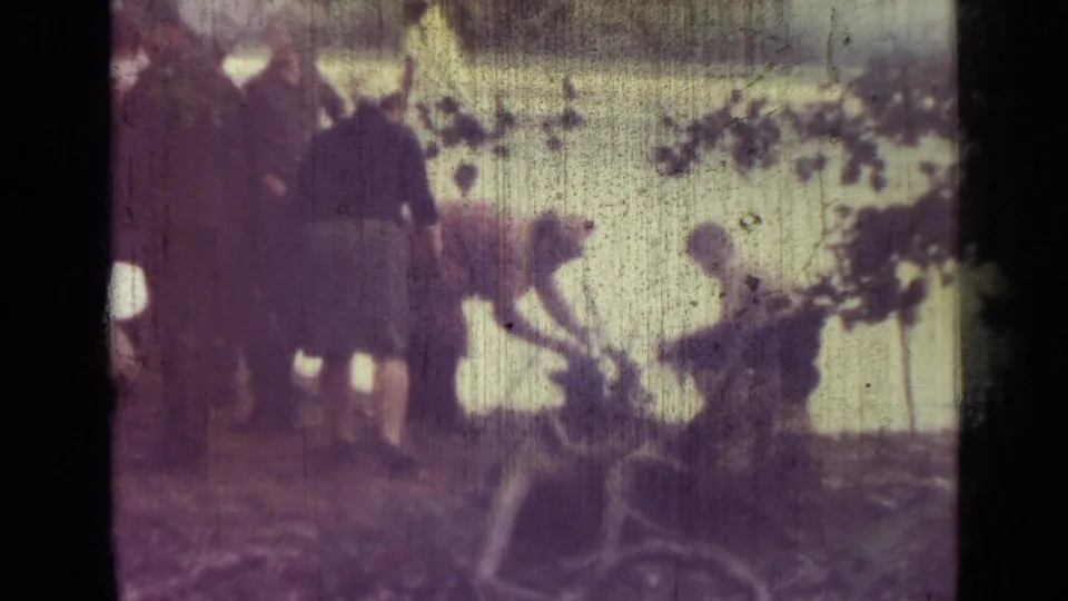 1967 8mm home-movie frame near Saint Petersburg showing adults beside a lake, with a tent, shoreline, and overhanging branches visible.