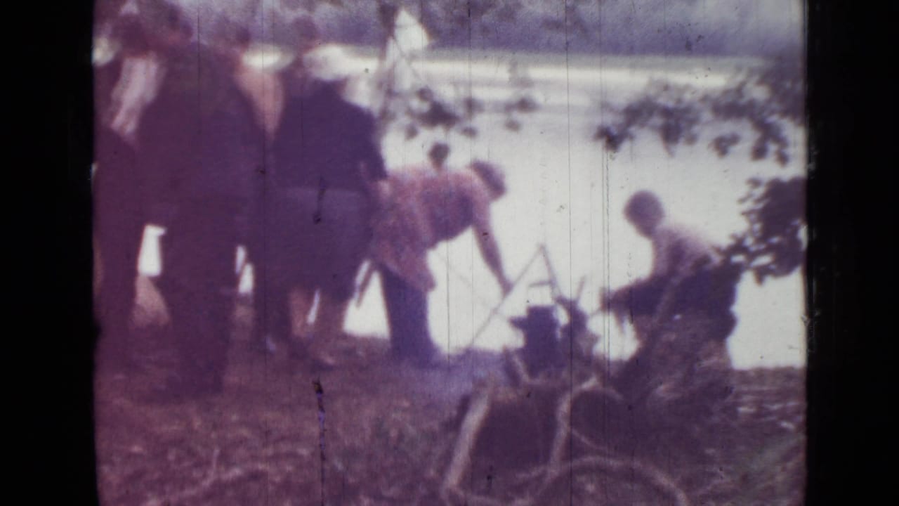 Campers stand and bend near the lake while gear sits on the ground by the shore.