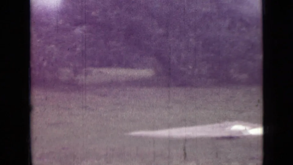 1967 8mm home-movie frame from the Saint Petersburg metadata showing a grassy clearing, pale camping fabric, and a wooded backdrop.