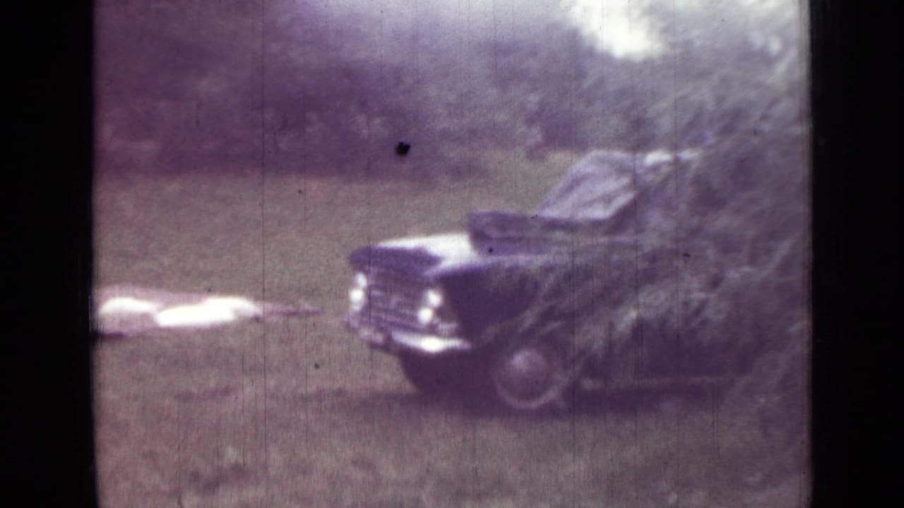 1967 8mm home-movie frame from the Saint Petersburg metadata showing a parked car partly obscured by trees near camping fabric.