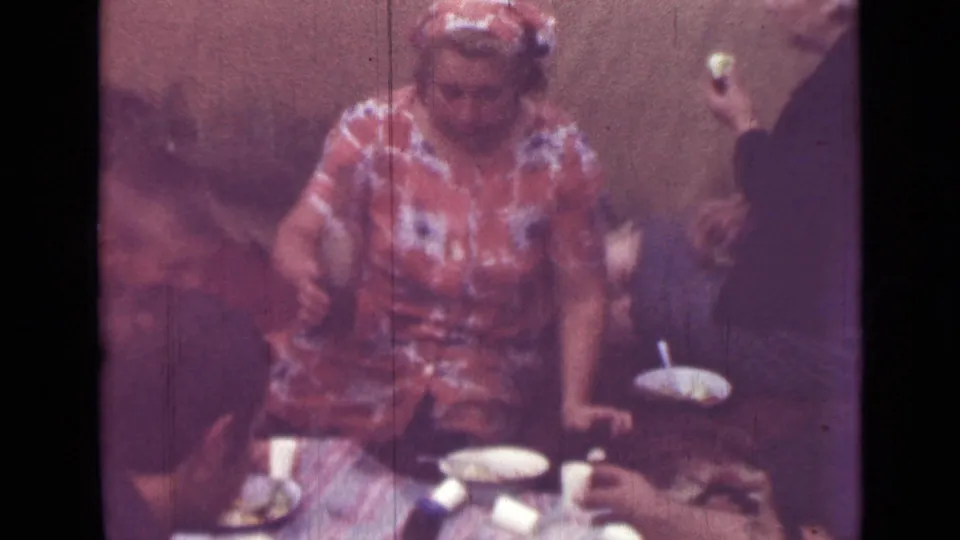 1967 Saint Petersburg home-movie frame showing a woman in a red patterned dress and headscarf serving food at an indoor gathering.