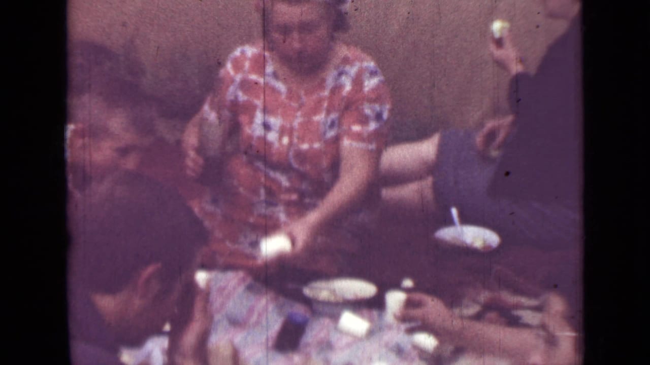 People gathered around a small table while a woman in a patterned dress handles food or drink.