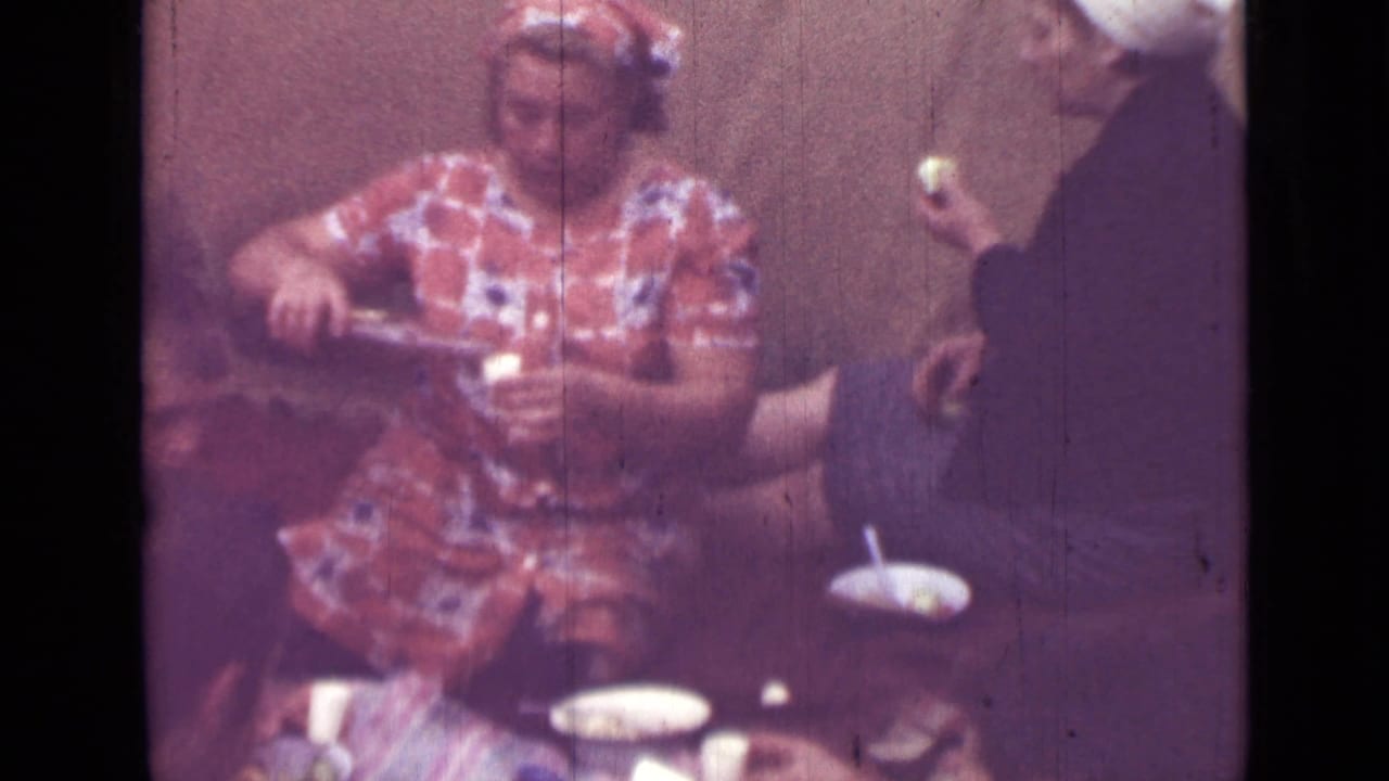 Woman seated at a table with cups and plates while another person stands nearby.