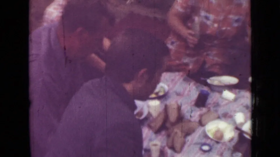 1967 Saint Petersburg home-movie frame with a seated man leaning toward food and dishes as others stand around the picnic blanket.