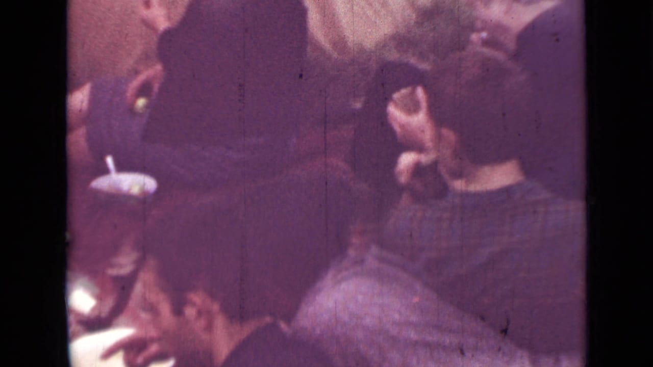1967 Saint Petersburg home-movie frame with adults seated around a small table, one person holding a cup or dish near the meal.