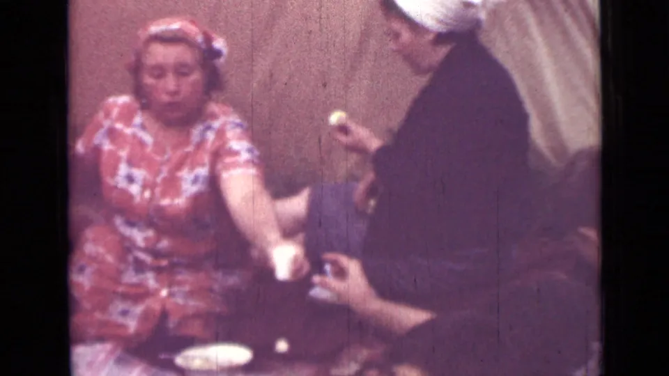 1967 Saint Petersburg home-movie frame showing a woman in a red patterned dress and headscarf seated at a meal as others pass food.