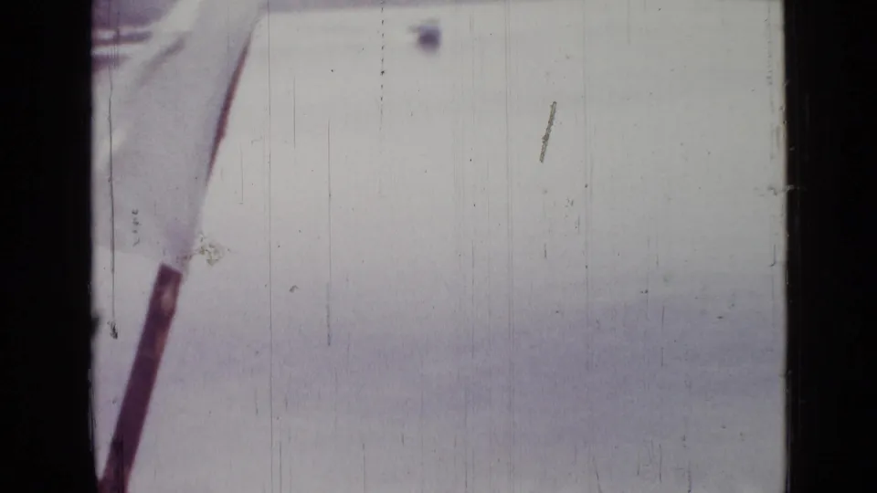 Soft 8mm frame of a pale lakeside view, with a dark diagonal pole or boat edge near the left and no clearly visible people.