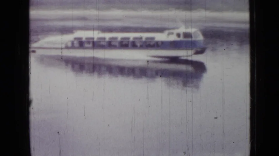Closer 8mm view of the white-and-blue vessel, showing its window row and curved bow as it moves across calm water.
