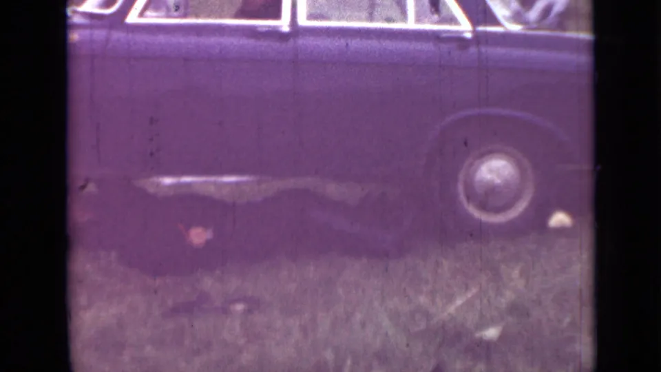 Dark sedan in a grassy field in 1967 Saint Petersburg, apparently raised for underside work, with rear wheel and soft 8mm texture visible.